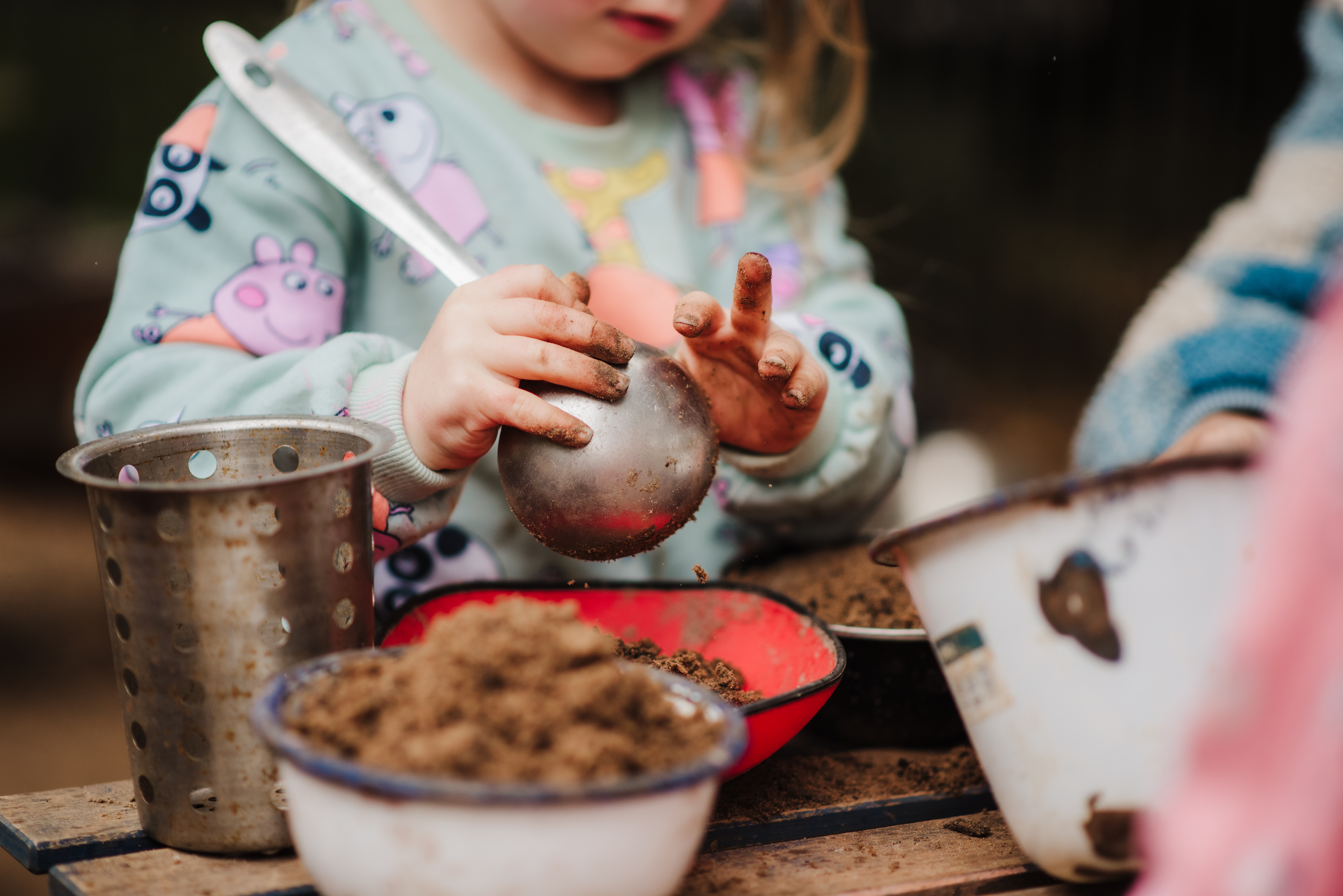 Child in jumper playing in a mud kitchen with a ladel and dirt-filled metal bowls