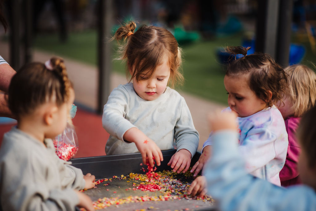 Young children with hands in, playing with coloured paper in a sensory bin.