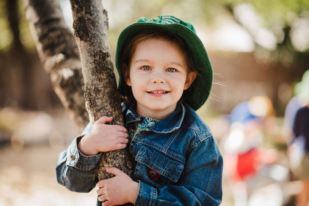 Girl wearing a blue denim jacket and a green hat, hugging a tree branch