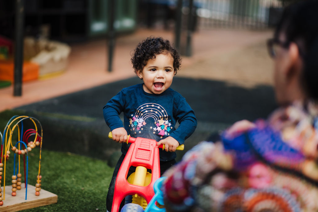 A curly-haired toddler riding on a toy bike with a big smile on her face