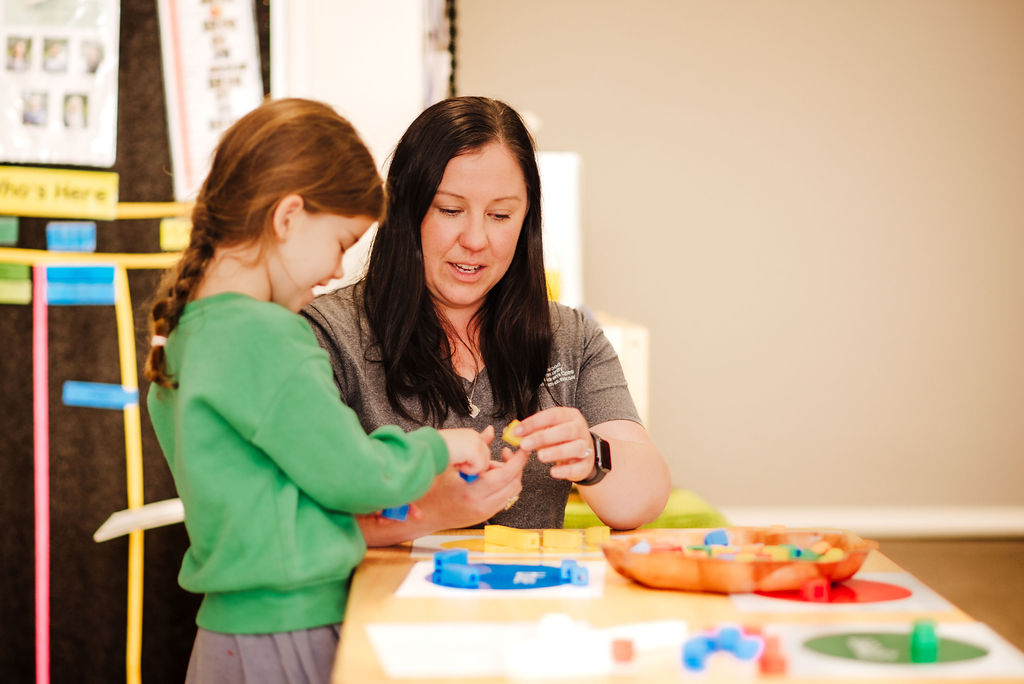 An educator with long brown hair with a child in a green top looking at small blocks on a table