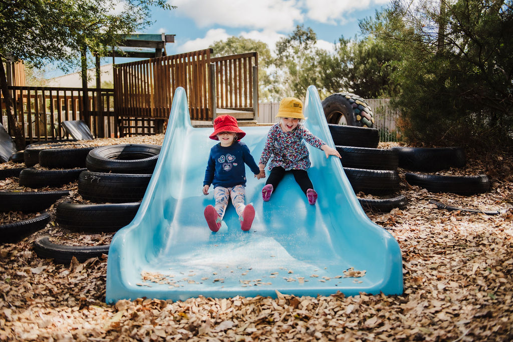 Two children on a big light blue slide, holdings hands and looking happy